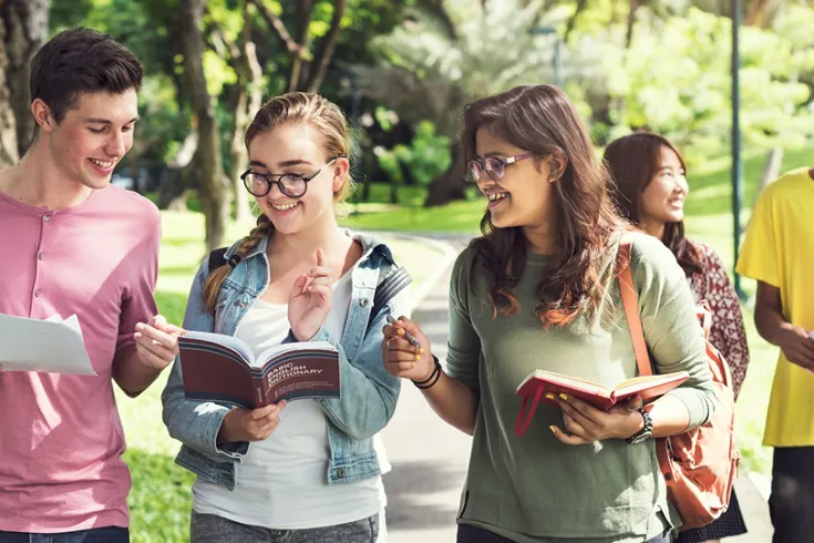 Three students walking in a park in the sun looking at books and smiling and talking