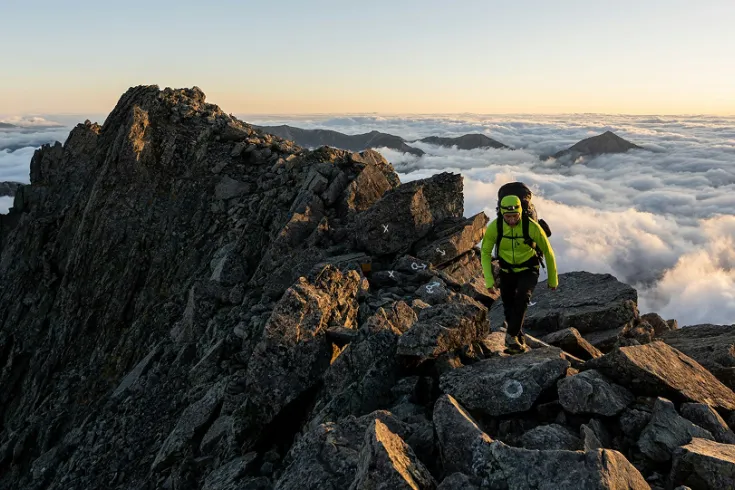 A hiker walking atop a mountain with clouds below