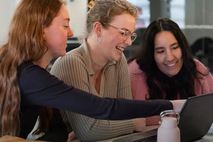Three women sat at a table looking at laptop and smiling