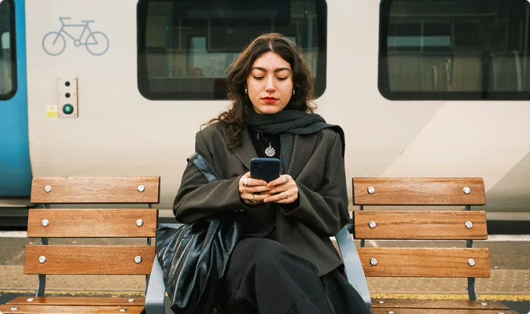 Woman sitting on a bench at a train station. She is looking at a phone in her hands and there is a train on the platform behind her.