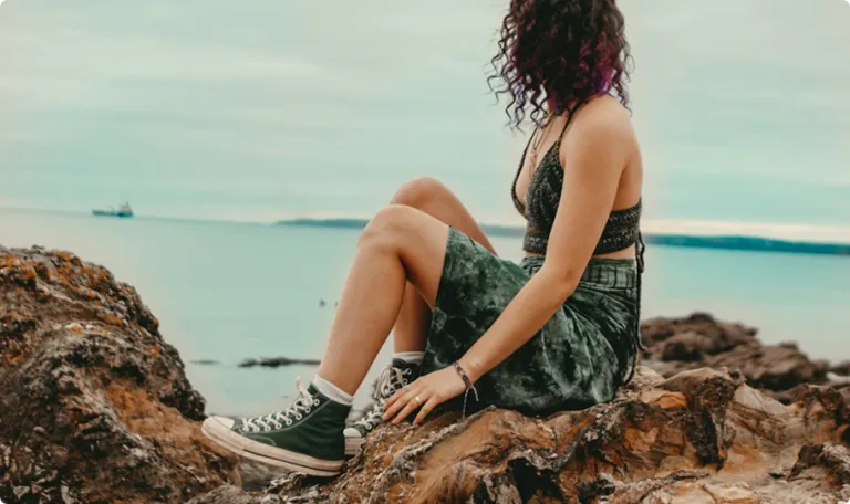 Woman sitting on some rocks in a coastal area, the ocean is in the background and she is looking out to sea.
