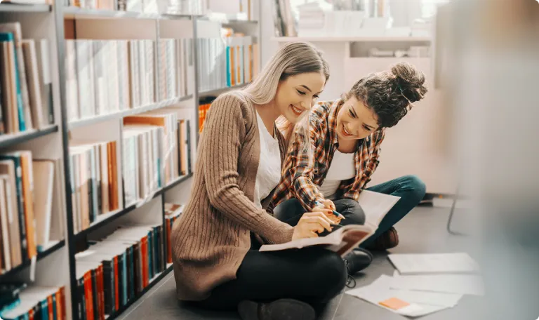 Two student sitting on the floor in a library writing in notebooks and smiling