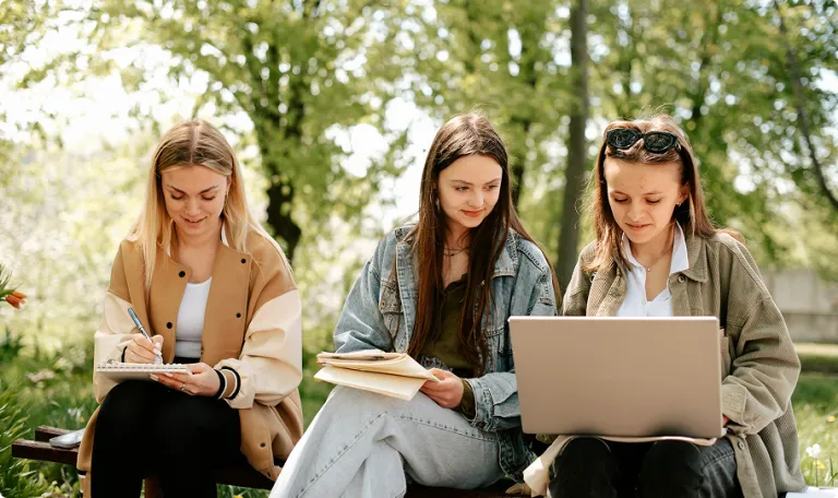 Three women sat in a tree lined park talking and looking at a laptop and notebooks