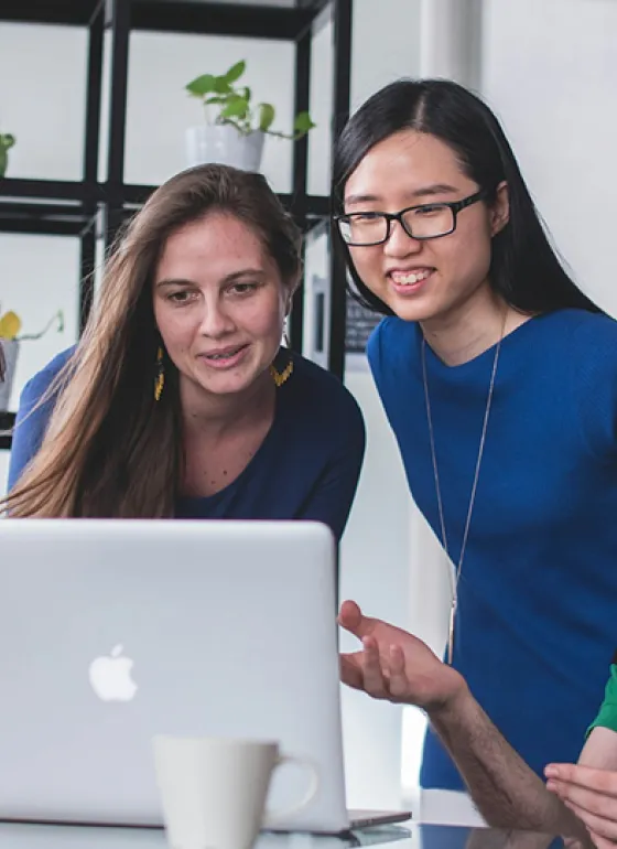 Four students in a classroom gathered around a laptop looking at the screen and talking to each other